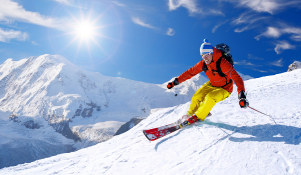 Man in red jacket and yellow snow pants demonstrating a carve on a large snowy mountain.